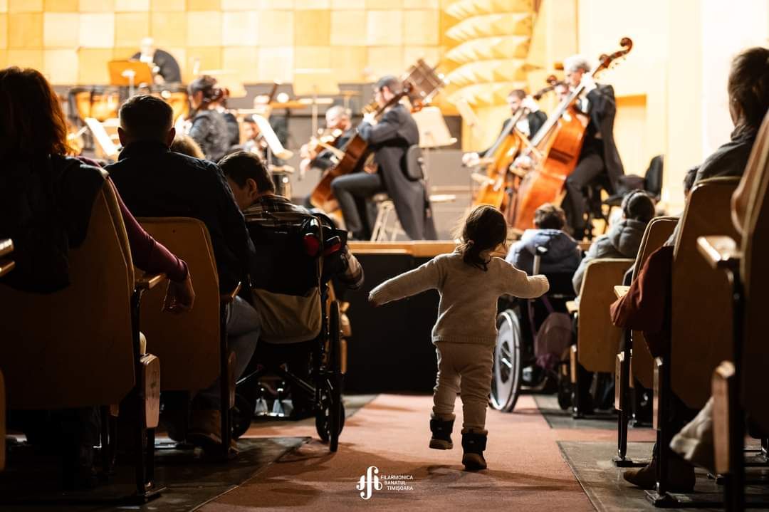 Concert la filarmonica Banatul unde un copil aleargă între rânduri spre scenă. În fața scenei se află persoane în scaune cu rotile.